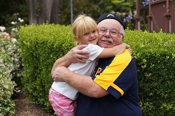 A man with glasses and a backwards baseball cap hugs his granddaughter outside